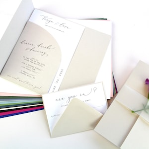 May include: A wedding invitation suite featuring a cream-colored pocket folder with a curved cutout, displaying text that reads Tanya & Levi are getting married, dinner, drinks & dancing, and June 25 2022. Includes a matching RSVP card with the text are you in?