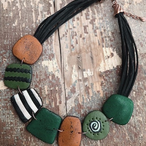 Könnte beinhalten: Handgefertigte Halskette mit geometrischen Formen in Grün, Braun und Weiß, auf mehreren schwarzen Kordeln aufgereiht. Die Halskette hat einen kupferfarbenen Verschluss und Verbinder.