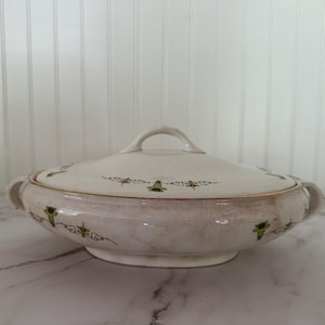 May include: Cream-coloured, oval-shaped serving dish with a lid. The dish has a handle on the lid and two side handles. It is decorated with green and gold floral patterns. The dish is on a white and grey marble surface.