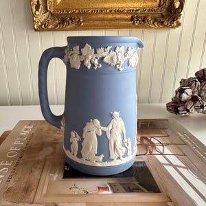May include: A blue ceramic pitcher with a handle and a decorative white relief design. The pitcher features a grape vine design around the top and classical figures around the body. The pitcher is sitting on a book.