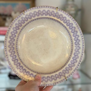 May include: A vintage cream-colored plate with a decorative border in a light purple hue. The plate is held by a gold-colored stand, showcasing its design. The border features a repeating pattern of connected rectangles and small floral elements.