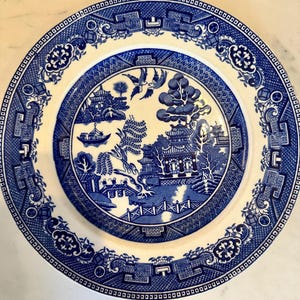 May include: A blue and white ceramic plate featuring a classic Willow pattern design. The plate has a detailed border and a central scene depicting a bridge, buildings, and trees. The plate is round.