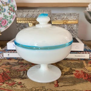 May include: A white glass candy dish with a lid, featuring a turquoise blue accent band and a matching finial. The dish is elevated on a pedestal base. Decorative home decor.