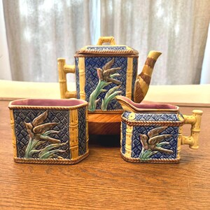May include: A ceramic tea set with a teapot, creamer, and sugar bowl. The set features a blue and yellow bamboo design with bird and plant relief details. The teapot has a striped spout and handle. The sugar bowl and creamer have a pink interior.