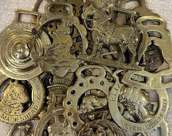 May include: A collection of vintage brass horse brasses featuring various embossed designs including a ship, a horse, a dog, and a profile of Queen Elizabeth II with the text Silver Jubilee 1977. These decorative metal ornaments have loops.