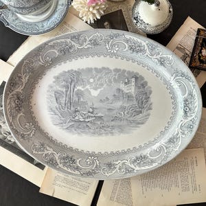 May include: An oval ceramic serving platter featuring a gray and white transferware pattern. The center depicts a landscape with a castle, boats on water, and a moonlit sky. The wide border displays intricate floral scrolls and geometric motifs.