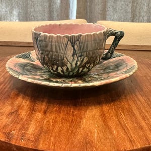 May include: A vintage teacup and saucer set. The cup and saucer are decorated with a textured, leaf-like pattern in shades of gray, green, and pink. The interior of the cup is pink. The set is placed on a wooden surface.
