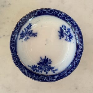May include: A small, round, antique white and blue dish with a floral design. The rim is a deep blue with a gold trim. The center of the dish is white with three clusters of blue floral patterns.