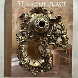 May include: Ornate, antique brass fixture with intricate floral and scroll detailing. The fixture is resting on a book titled "SENSE OF PLACE" with text that reads "DESIGN IN BY WHERE WE LIVE". The fixture has a central hole and a decorative, sunburst-like design.