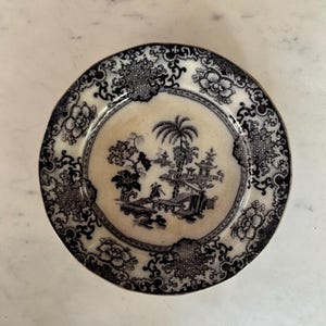 May include: An antique plate with a black and white Asian-inspired design. The centre features a landscape with a palm tree, figures, and a pagoda. The rim has floral and geometric patterns.