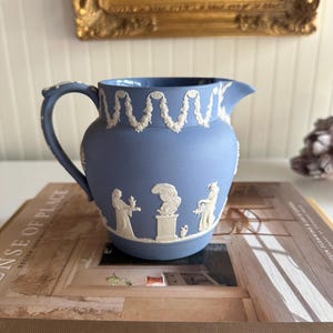 May include: A blue ceramic pitcher with white relief decorations. The pitcher features a handle, spout, and a detailed design of classical figures and floral garlands. It is resting on a book with the title "SENSE OF PLACE".