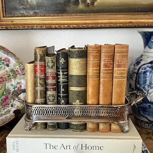 Può includere: Un cestino in metallo argentato con manici e piedini decorati, contenente una fila di sette libri antichi con dorsi in pelle e tessuto vari. Il cestino poggia su due libri con copertina rigida impilati intitolati The Art of Home e A Year in the French Style.
