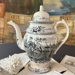 May include: A cream-colored ceramic teapot featuring a black transferware pattern depicting a coastal landscape with figures working in a field, sailboats on the water, and floral borders. The teapot includes a matching lid with a finial top.