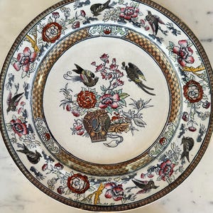 May include: A set of decorative plates with a floral and bird design. The plates feature a cream-colored background with a brown and gold border. The center of the plates has a vase with flowers and birds. The plates are likely made of ceramic.