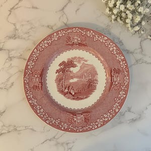 May include: A round, antique-style plate with a red and white landscape design. The plate features a central scene of a tree, figures, and a building, surrounded by a decorative border. The plate is likely made of ceramic or porcelain.