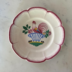 May include: A white ceramic plate with a scalloped edge and a burgundy trim. The plate features a colorful rooster design in the center, with a blue and white basket and green foliage. The plate is likely for decorative or serving purposes.