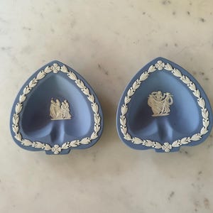 May include: Two heart-shaped, light blue ashtrays with white relief detailing. Each ashtray features a white relief design of figures and a white floral and leaf border. The ashtrays are displayed on a marble surface.