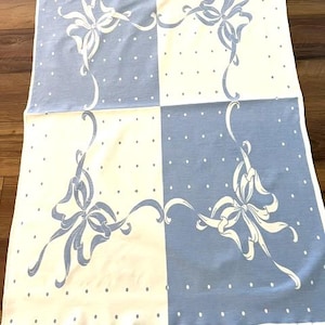May include: A rectangular textile featuring a checkerboard pattern of light blue and white squares. Each square is decorated with a white or light blue ribbon bow motif and a repeating pattern of small dots. The edges have a scalloped finish.