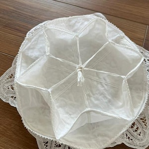 May include: A white fabric bowl cover featuring a star-shaped interior structure with six segments meeting at a central knotted point. The circular cover is edged with a decorative white lace trim that extends outward in a scalloped pattern.