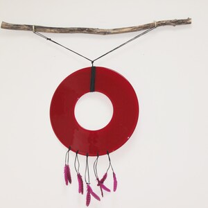 May include: A red circular glass piece with a hole in the center hangs from a branch with black string.  Purple feathers dangle from the bottom of the glass piece.