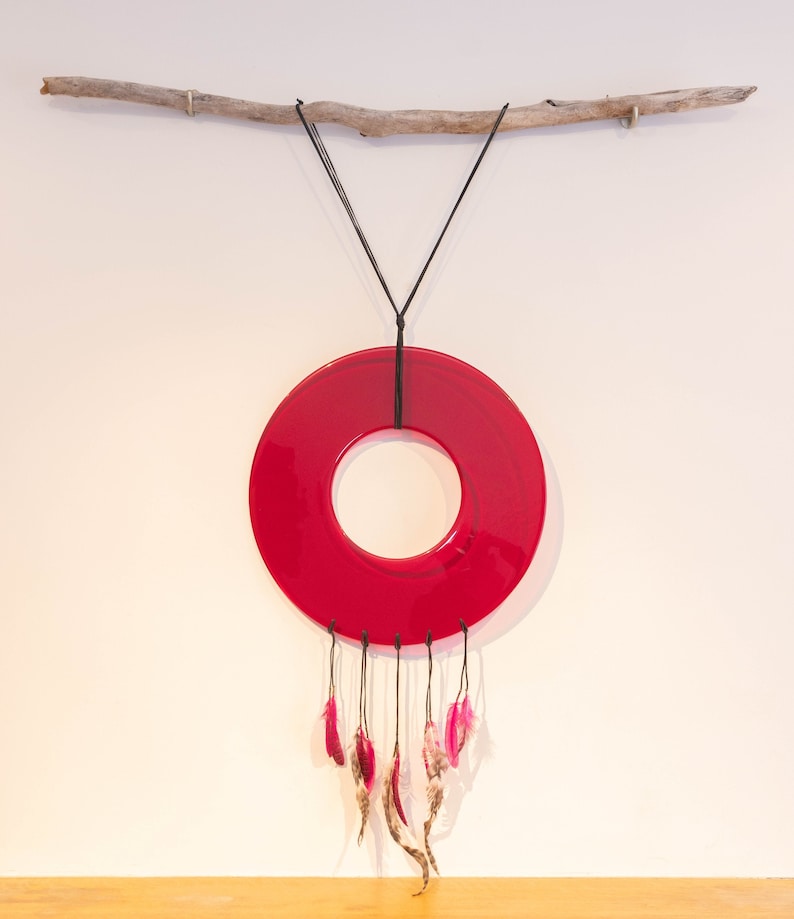 May include: A red glass circle with a hole in the center hangs from a branch with black string.  Pink and brown feathers dangle from the bottom of the circle.