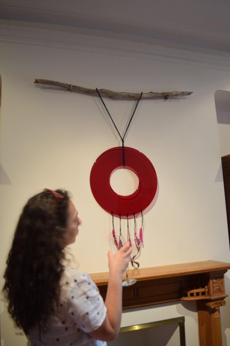 May include: A red circular glass mobile hangs from a branch attached to the wall. The mobile has a ring of pink glass pieces hanging from it.