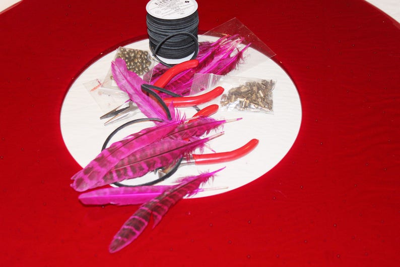 May include: A set of pink feathers, black elastic, pliers, and gold beads on a white surface. The items are arranged in a circle, with the feathers in the center.