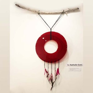 May include: A red glass circle with a hole in the center hangs from a branch. Pink feathers dangle from the bottom of the circle. The text "By Nathalie Guetz" is written on a white label below the circle.