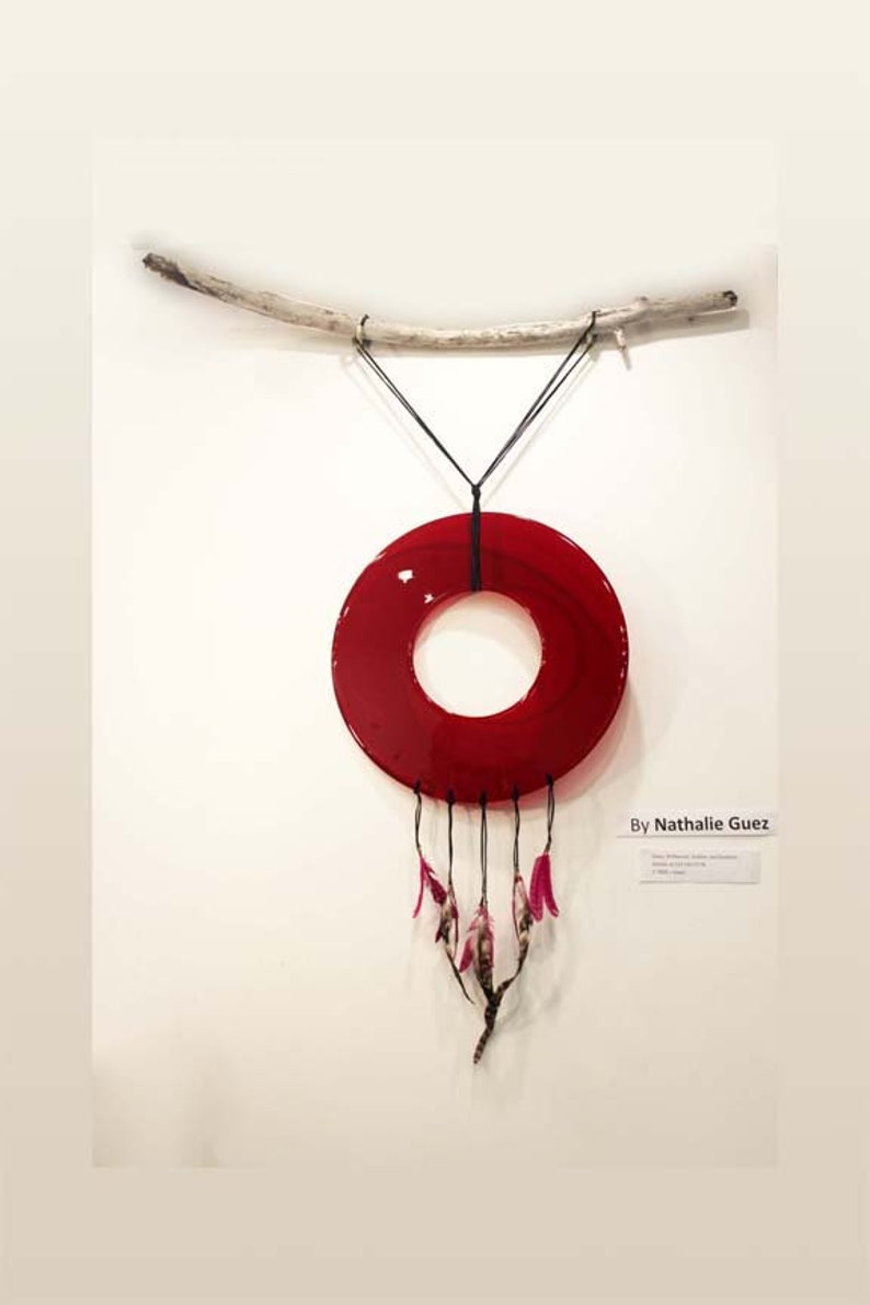 May include: A red glass circle with a hole in the center hangs from a branch. Pink feathers dangle from the bottom of the circle. The text "By Nathalie Guetz" is written on a white label below the circle.