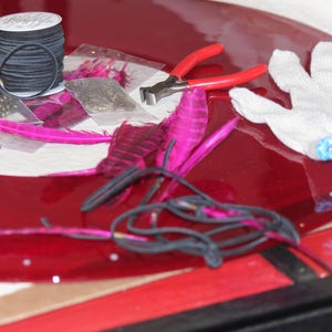 May include: A red vinyl record with a variety of craft supplies on top, including pink feathers, black thread, beads, and a pair of pliers. A white cotton glove is also on the record.