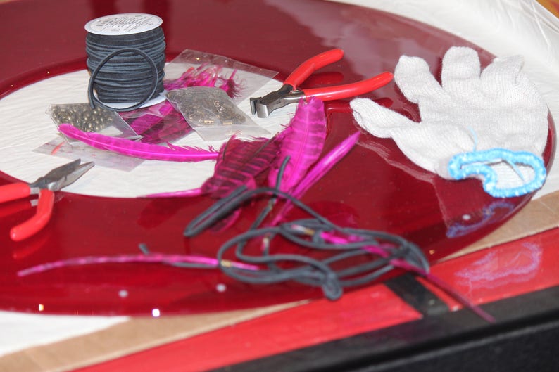 May include: A red vinyl record with a variety of craft supplies on top, including pink feathers, black thread, beads, and a pair of pliers. A white cotton glove is also on the record.