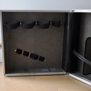 May include: Open silver case with black straps and a gray fabric interior. The case has a black leather strap and a gray fabric pocket.