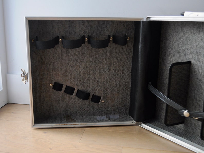 May include: Open silver case with black straps and a gray fabric interior. The case has a black leather strap and a gray fabric pocket.