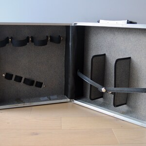 May include: An open silver aluminum case with a gray fabric interior. The case has multiple compartments and straps for holding items.