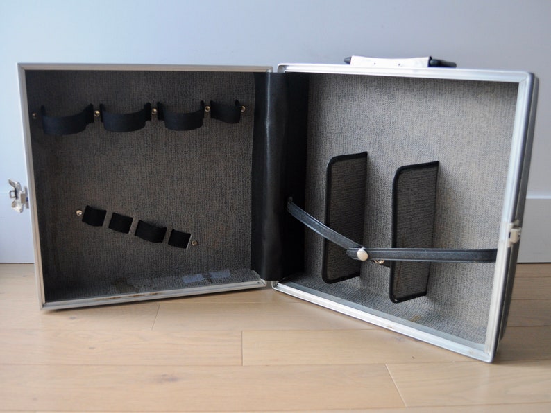 May include: An open silver aluminum case with a gray fabric interior. The case has multiple compartments and straps for holding items.