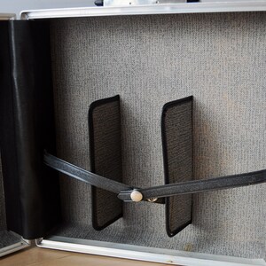 May include: The inside of a silver aluminum case with a gray fabric lining. Two black fabric straps are attached to the case with a metal buckle. The straps are used to secure items inside the case.