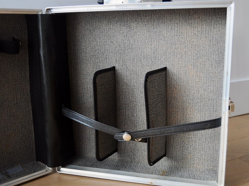 May include: The inside of a silver aluminum case with a gray fabric lining. Two black fabric straps are attached to the case with a metal buckle. The straps are used to secure items inside the case.