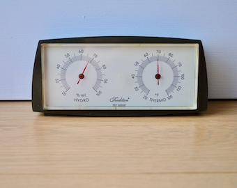 May include: A black and white analog hygrometer and thermometer with a red needle on each gauge. The gauges are labeled "% rel. HYGRO" and "°F THERMO 120". The device is made by Tradition, West Germany.