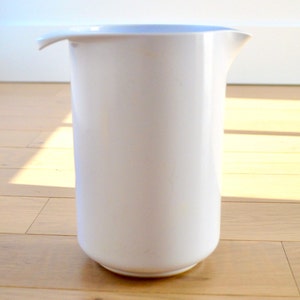 May include: A white, cylindrical jug with a curved spout and a rounded base. The jug is made of a smooth, glossy material and sits on a light-coloured wooden floor. The background includes a white wall and a window.