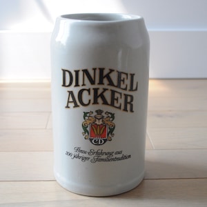 May include: White ceramic beer stein with a black and gold logo that reads "Dinkel Acker" and the text "Brau-Erfahrung aus 200 jähriger Familientradition".