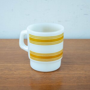 Può includere: Tazza in ceramica bianca con un motivo a strisce dorate. La tazza ha un manico ed è appoggiata su una superficie di legno.