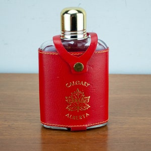 Pode incluir: Frasco de couro vermelho com tampa de metal dourado e uma inscrição dourada que diz "Calgary Alberta" com um design de folha de bordo dourada.