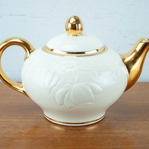 Könnte beinhalten: Weiße Teekanne aus Keramik mit goldenen Verzierungen und einem floralen Design. Die Teekanne hat einen Deckel mit einem goldenen Knopf.
