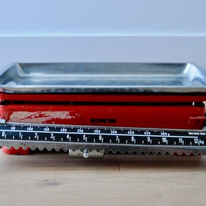 May include: A vintage red kitchen scale with a stainless steel tray. The scale has a black measuring tape with markings in grams and kilograms. The scale is labelled "EKS" and "Made in Sweden".