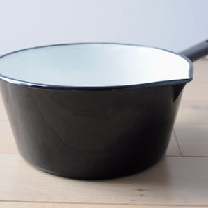 Op de afbeelding: Een zwarte emaille pan met een wit interieur en een zwarte handgreep. De pan staat op een licht houten oppervlak.