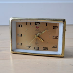 May include: A gold-colored alarm clock with a white face and black numbers. The clock has a gold-colored frame and a red heart logo with the text "Five Home" on the face.