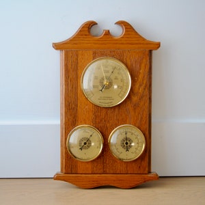 May include: A wooden wall-mounted barometer with three round dials. The top dial is labeled "The Old English Barometer" and the bottom two dials are labeled with the words "Humidity" and "Temperature".
