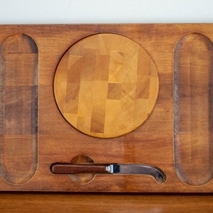 May include: A wooden cheese board with a round cutting surface, two long rectangular grooves, and a small knife slot. The board is made of light brown wood and has a natural finish.