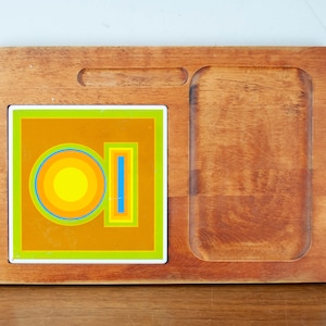 May include: A wooden serving tray with a removable tile featuring a geometric design in shades of yellow, orange, blue, and green. The tile is set into a recessed area of the tray.