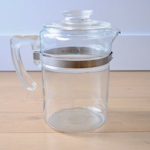 May include: A clear glass coffee pot with a metal band around the middle and a glass lid. The pot has a handle and a spout.